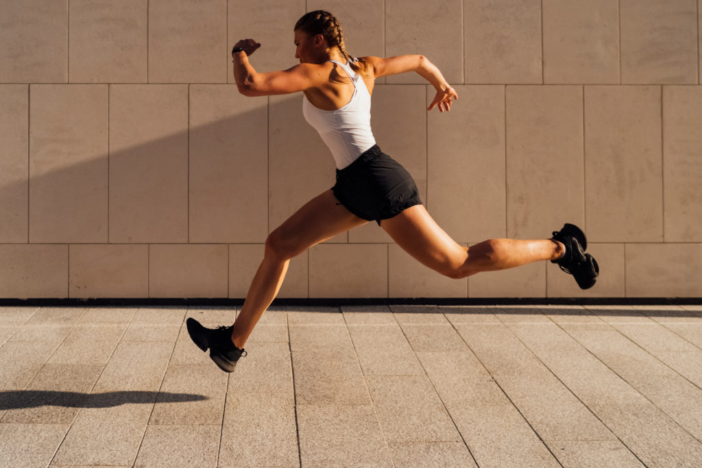 A young adult Caucasian woman is running outdoor crossing modern buildings exterior at sunset.