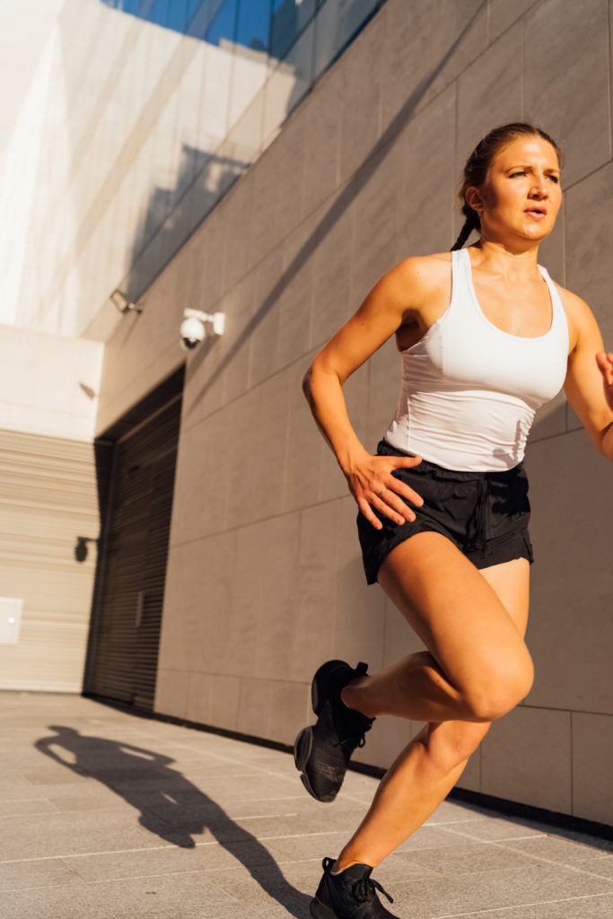 A young adult Caucasian woman is running outdoor crossing modern buildings exterior at sunset.