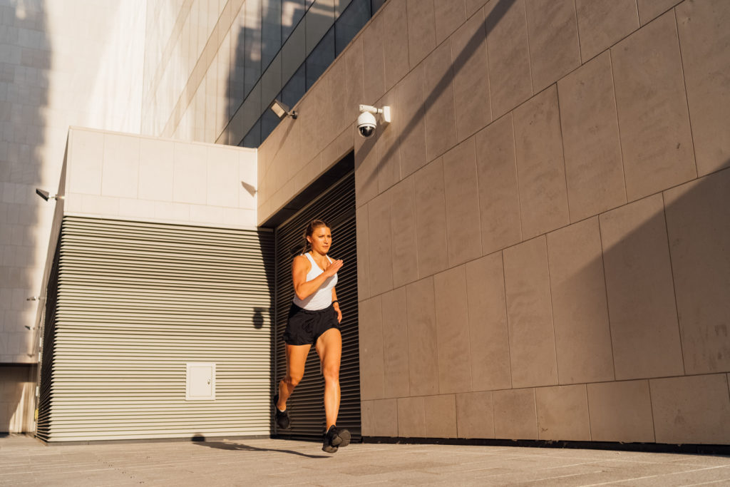 A young adult Caucasian woman is running outdoor crossing modern buildings exterior at sunset.