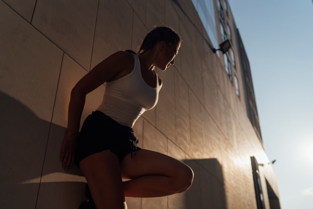 A Young Woman is stretching against a building wall in the city. The outdoor scene at sunset in backlight