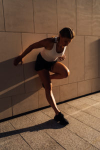 A Young Woman is stretching against a building wall in the city. The outdoor scene at sunset in backlight
