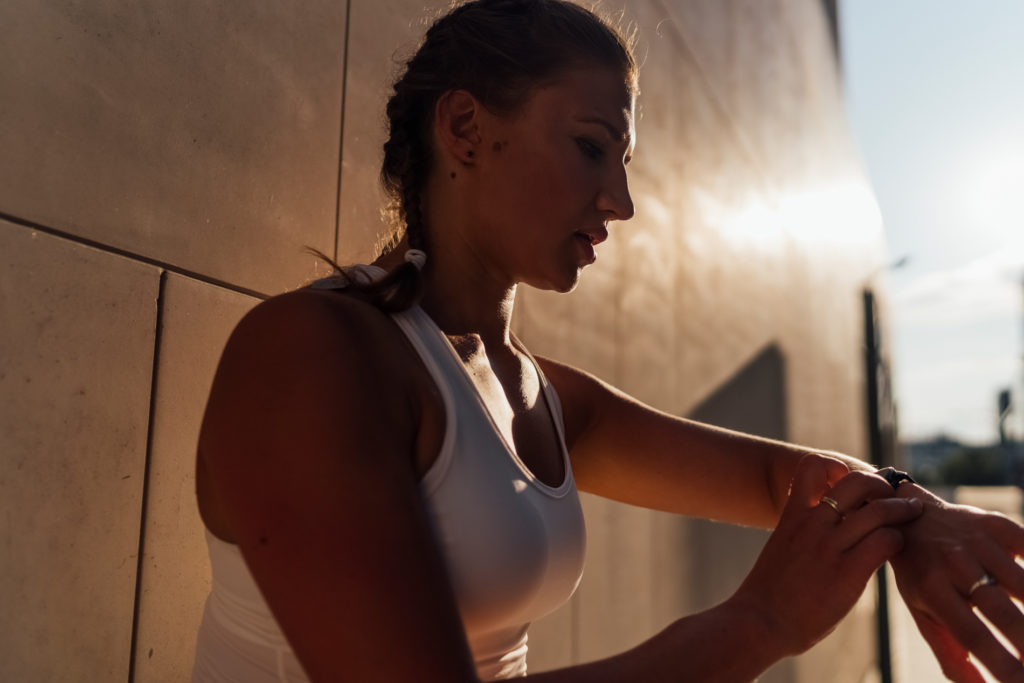 A young Caucasian woman is tapping the screen of the sports watch, looking for details of the sports activity against a modern wall. Outdoor scene at the sunset.