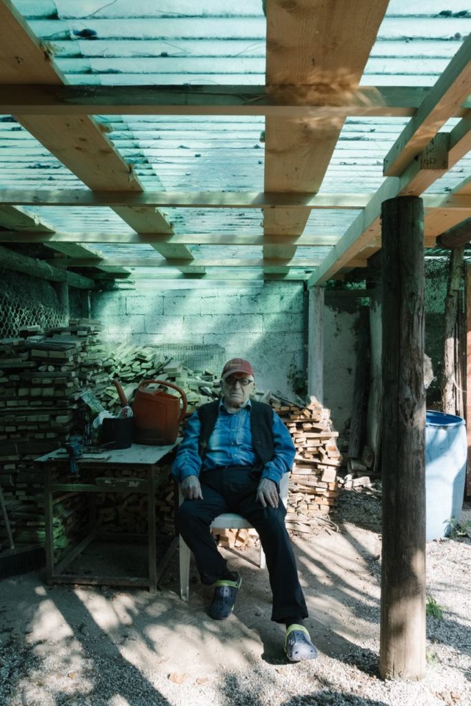 Old Caucasian Man Portrait seated under a green cover roof of a shack in the Backyard. Background full of wooden logs