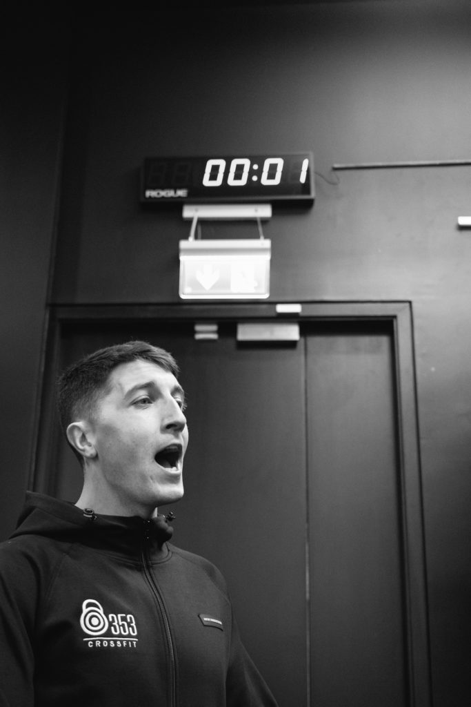 A Male Crossfit Coach counts down the Time During a Crossfit competition in the Gym. The clock on the top of the wall in the background and a Man screaming Loud in the foreground. Balck and white image