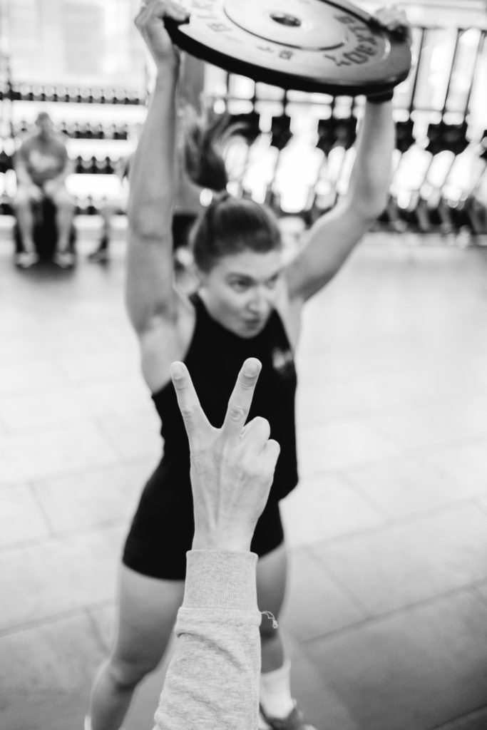 In the background of this Black and White Picture, a young woman is Lifting with energy a Disk Weight while an Anonymous Head, in the foreground, counts the number two of the repetitions. Cross Fit competition indoor