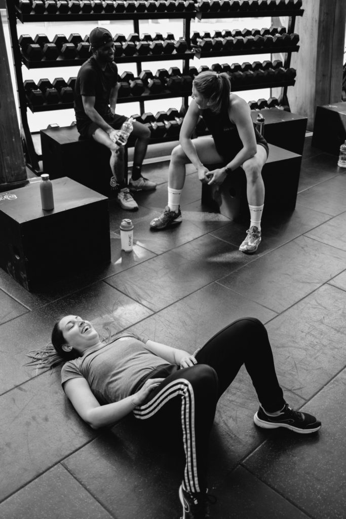 Portrait of a Tired woman Laughing on the Floor while the teammates are chatting in the background close to the Dumbell Rack. Black and White image
