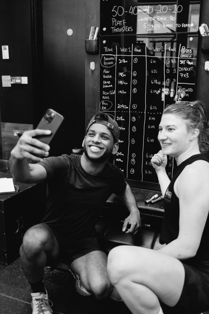 A black Young Man and a Caucasian Woman are taking a selfie in front of the Crossfit Gym's Board at the end of the Training