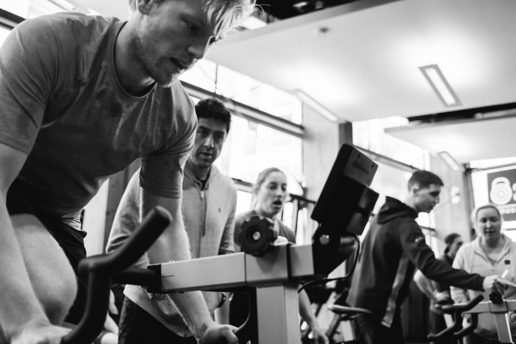 A young Caucasian Man is Sweating on a bike during a Crossfit Competition. More People in the background. Black and White image