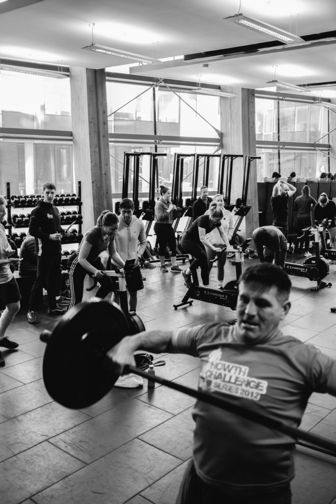 People Competing during a Crossfit Training. A real Caucasian Man in the Foreground is Lifting a Bar with Weights while more people in Focus in the Background are Exercising on a Bike. Black and White image