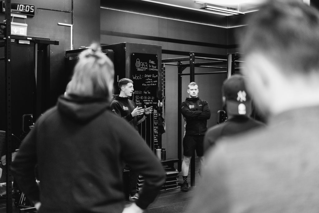 Two Coaches are talking in front of the Crossfit Board, Explaining how the Competition is developed. People out of focus Gathered in the Foreground. Black and White image