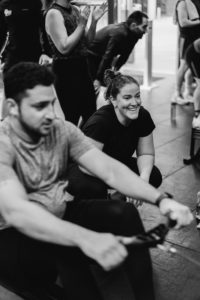 A caucasian Woman is Smiling and cheering Her Gym partner during a Crossfit competition in the Gym. The middle eastern Man is Rowing in the Row machine, and many people are in the foreground supporting each other. Back and white image.