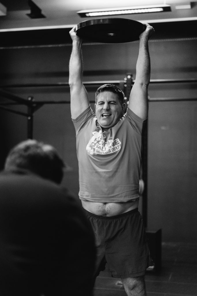 A Real Adult man is Lifting a Disk Weight during a CrossFit competition Training. Lifting His Arms, the Belly is visible. His team is waiting for his turn in the Foreground.