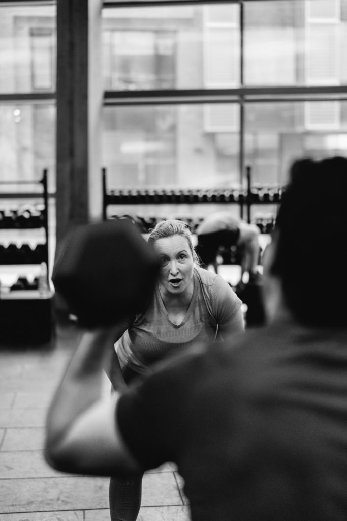 A real Caucasian Woman is Yelling and Cheering Her Teammate during a Crossfit competition in the Foreground, lifting a Dumbell.