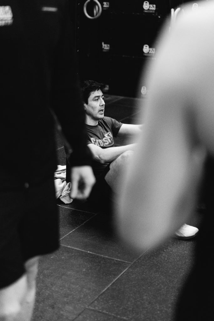 Tired Real Man is Seated on the Floor breathing Heavly at the Crossfit Gym. Two anonymous people are in the foreground. Black and White image