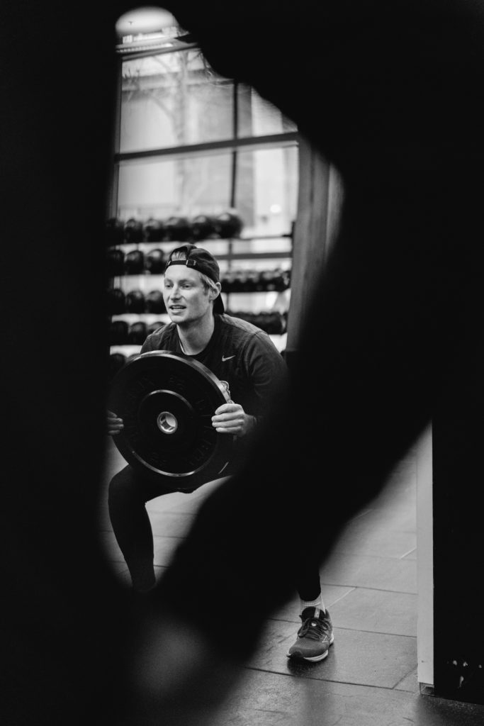An Adult Caucasian Man is squatting with a Weight Disk during a Crossfit Competition. The Man is Framed between the body and the arm of His Anonymous Teammate in the Foreground. Black and White image.