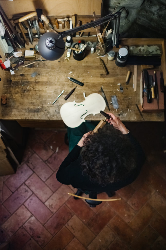 An Anonymous Italian Violinmaker sits on his work desk and works carefully on the part of a Handmade Violin.