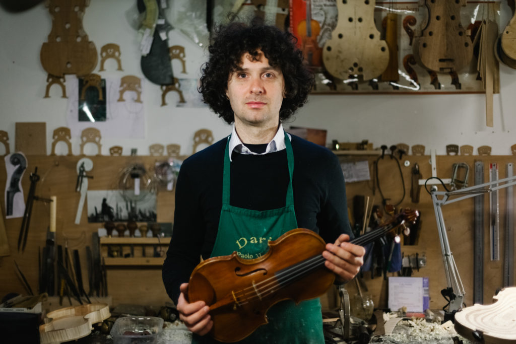 Italian Male Violinmaker portrait holding a Violin and Standing in front of his Workshop worktable