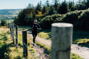 A trail runner moves along a sunlit forest path, surrounded by lush greenery and wooden posts. The image captures motion, energy, and connection to nature