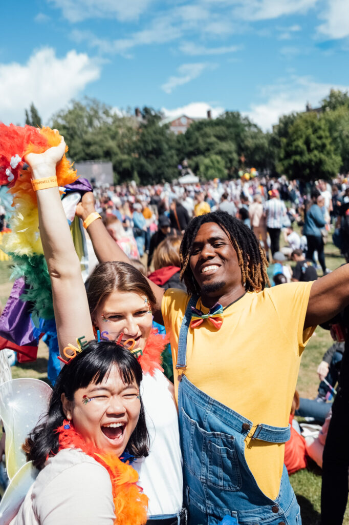 Three friends celebrating at Dublin Pride in a park, wearing colourful outfits and rainbow accessories.