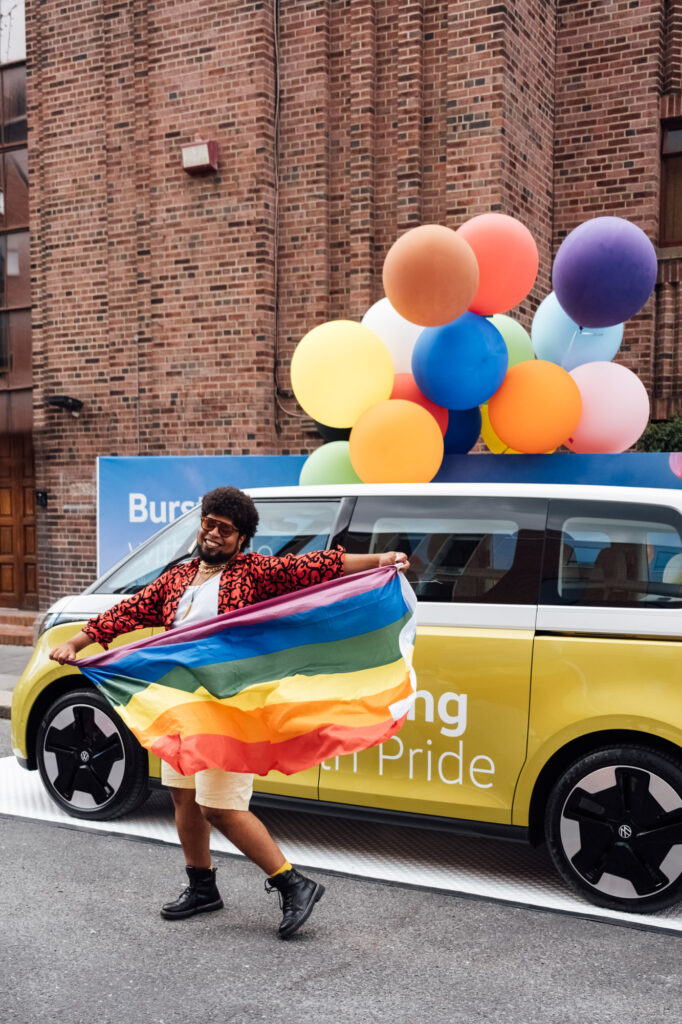 A joyful black man holding a rainbow flag in front of a Volkswagen ID. Buzz decorated with colourful balloons during Dublin Pride.