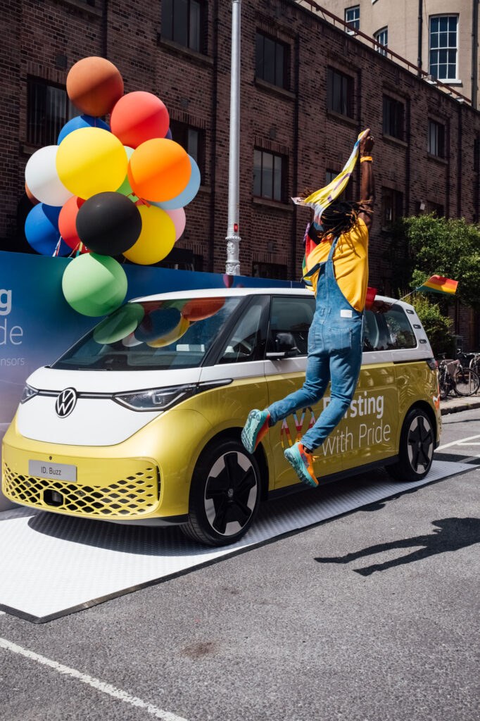 Person jumping in mid-air in front of a rainbow balloon-decorated VW ID. Buzz.