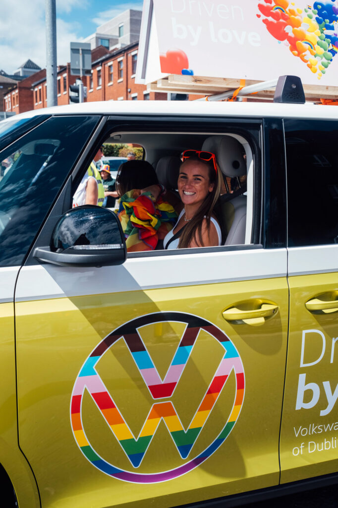 Smiling woman wearing red sunglasses seated in the driver’s seat of a yellow VW ID. Buzz decorated for Pride.
