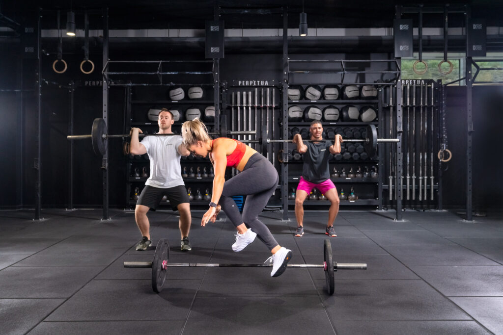 Athlete mid-air jumping over a barbell in a dynamic gym workout at Perpetua Dublin, with other athletes lifting in the background. Captured by Mattia Pelizzari Photography, specialising in sport and gym environments.