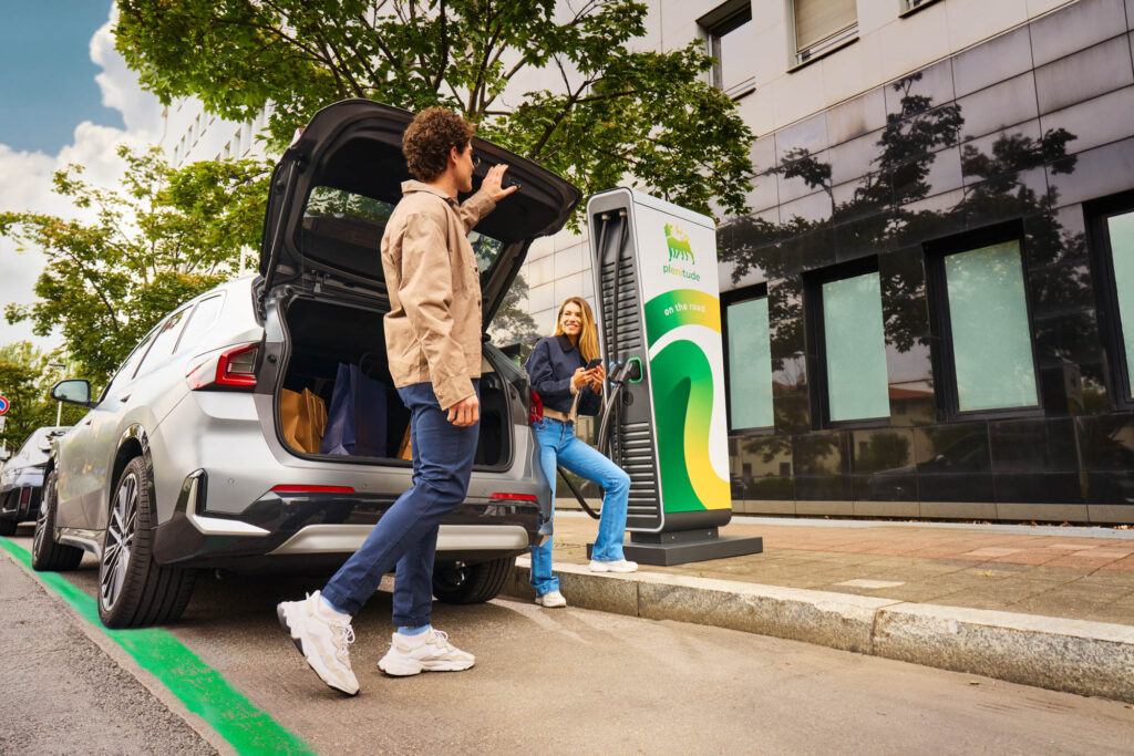 Man and woman by an electric car charging station, man closing the boot while woman unplugs the car smiling.