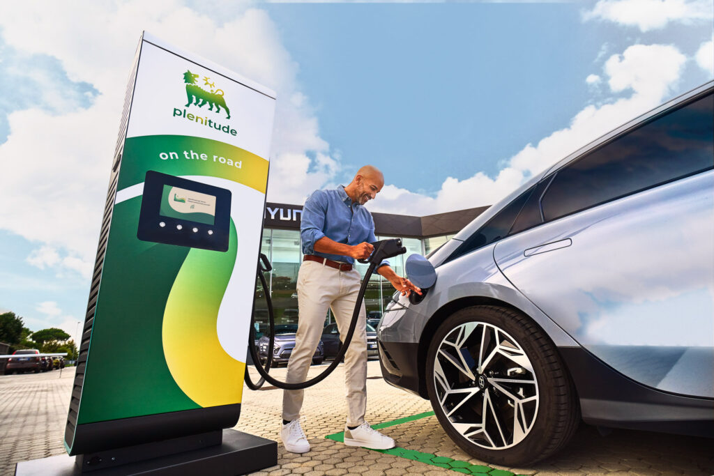 Man charging electric car at Plenitude ‘On the Road’ charging station under blue sky.