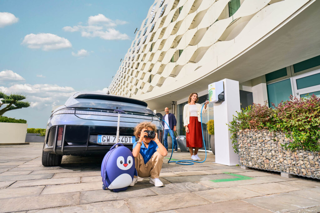 Family with electric car charging at Plenitude station, child squatting with camera in front