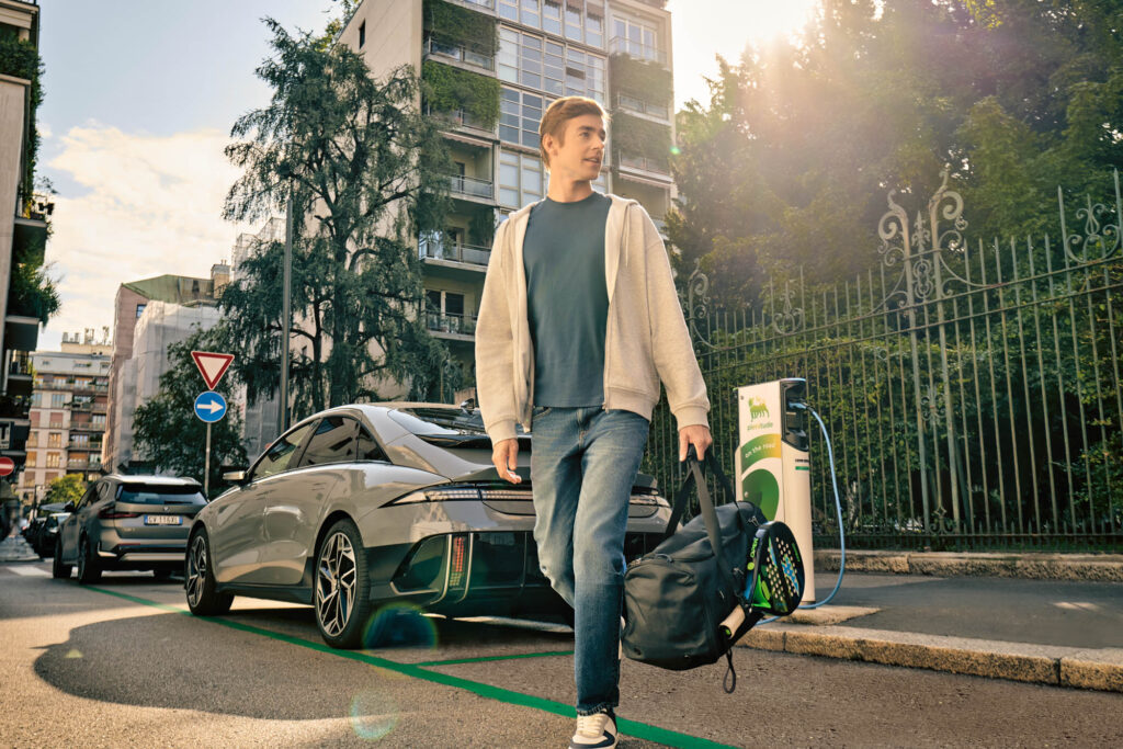 Young man walking away from his parked electric car while charging at a Plenitude charging station in an urban street.
