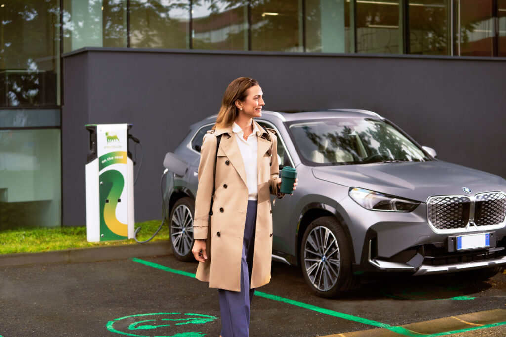 Woman walking with coffee cup past BMW charging at Plenitude electric vehicle station.