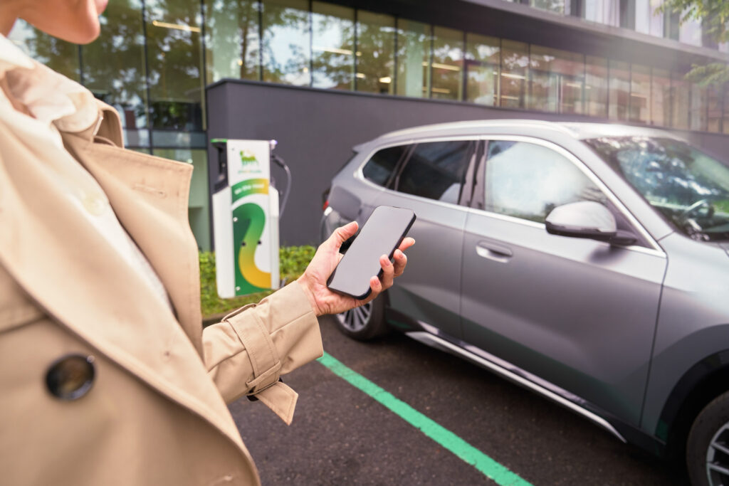 Person holding smartphone near silver BMW parked at Plenitude electric vehicle charging station.