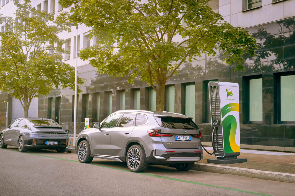 Two electric cars parked side by side and charging at Plenitude On the Road stations in a city street lined with trees.
